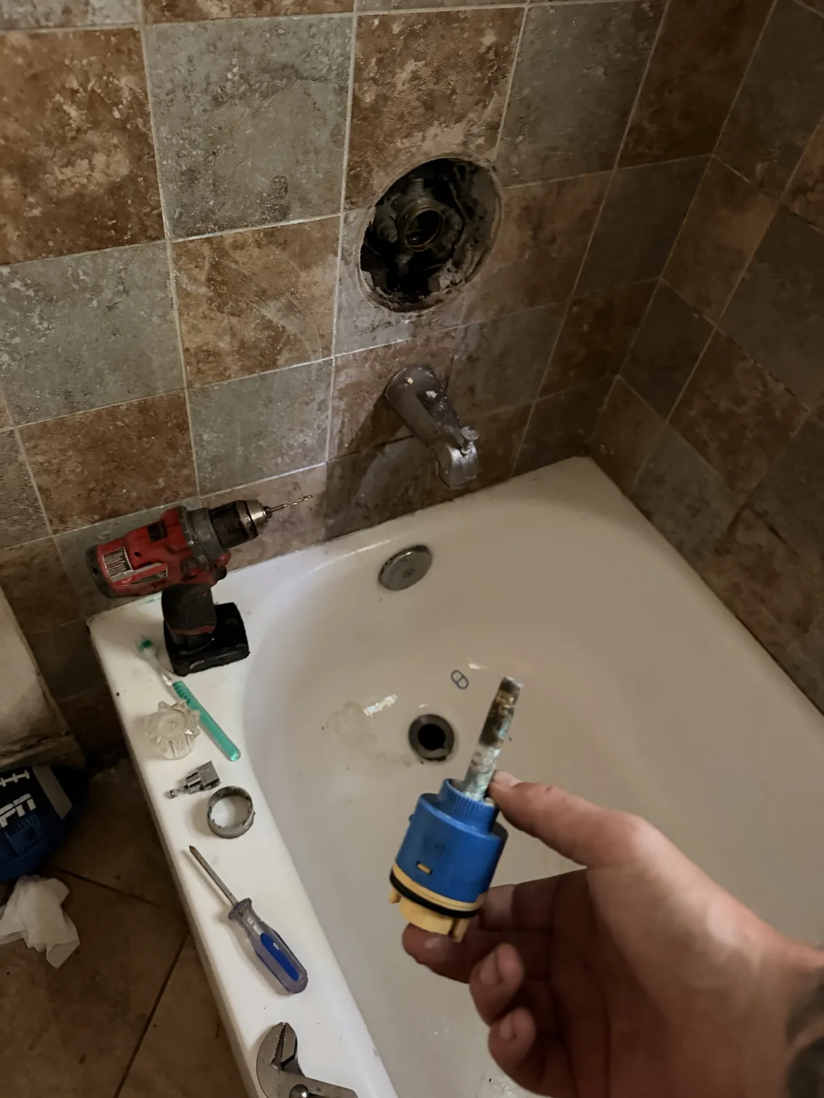 A shower valve cartridge held in a hand over a tub, with the open valve body in the tiled wall behind