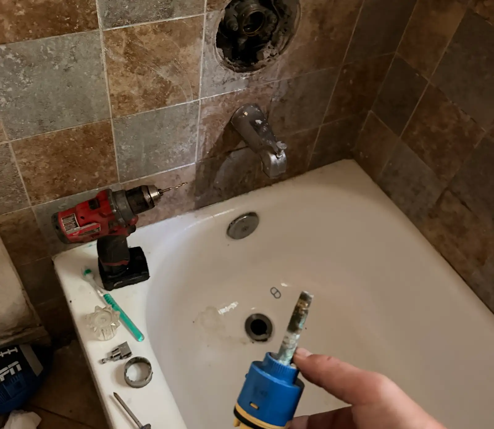 A shower and tub valve cartridge just removed and held up, its stem and seals furred with white mineral scale, the open valve body in the tiled wall behind it