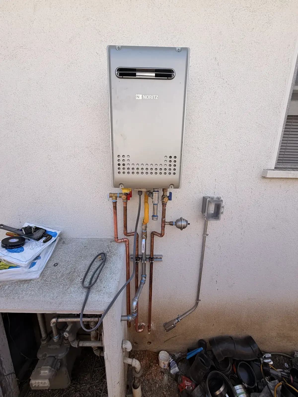 A wall-mounted tankless water heater on an exterior wall, with the gas and water connections made up and tools still on the bench below