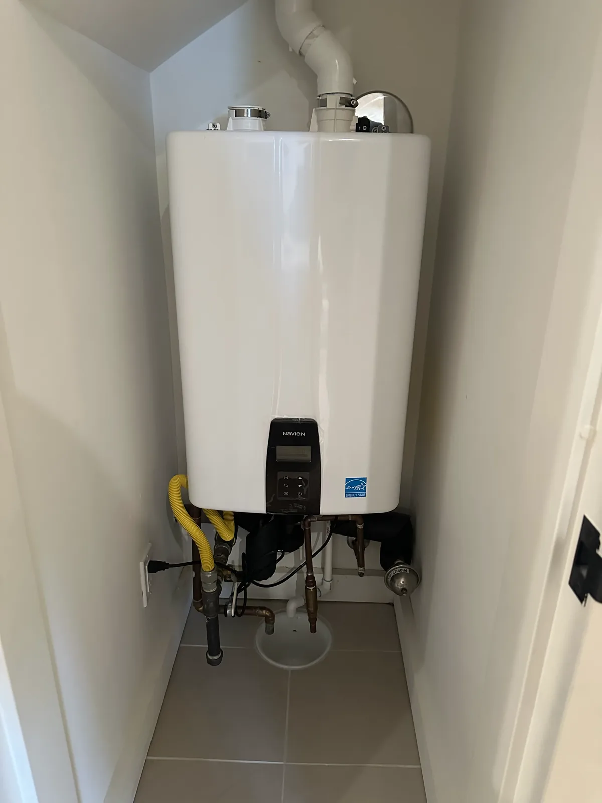 A tankless water heater mounted in a tiled interior closet with its venting and a yellow gas connector below