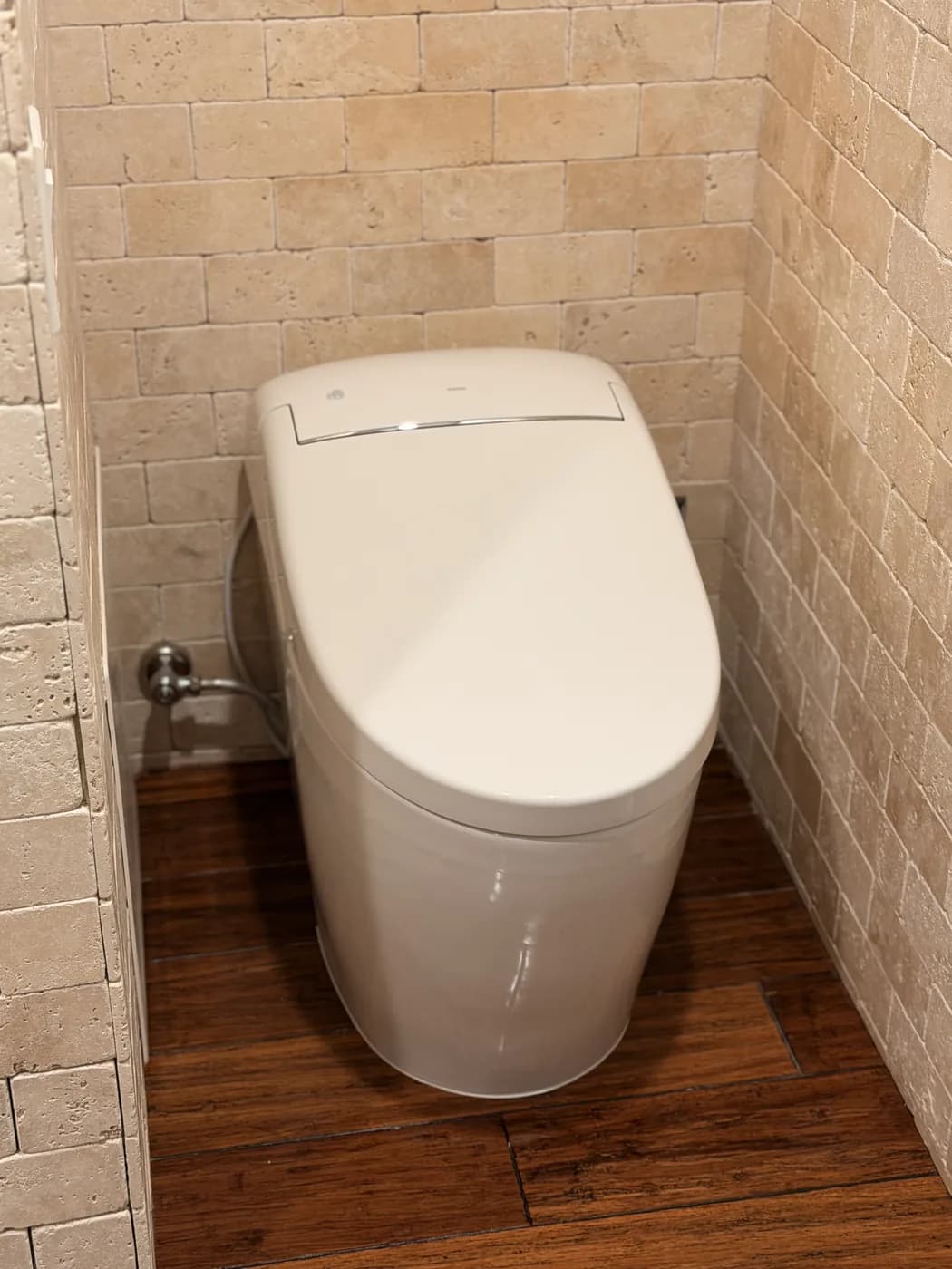 A one-piece skirted toilet installed in a narrow water closet with travertine-tiled walls and a dark wood floor
