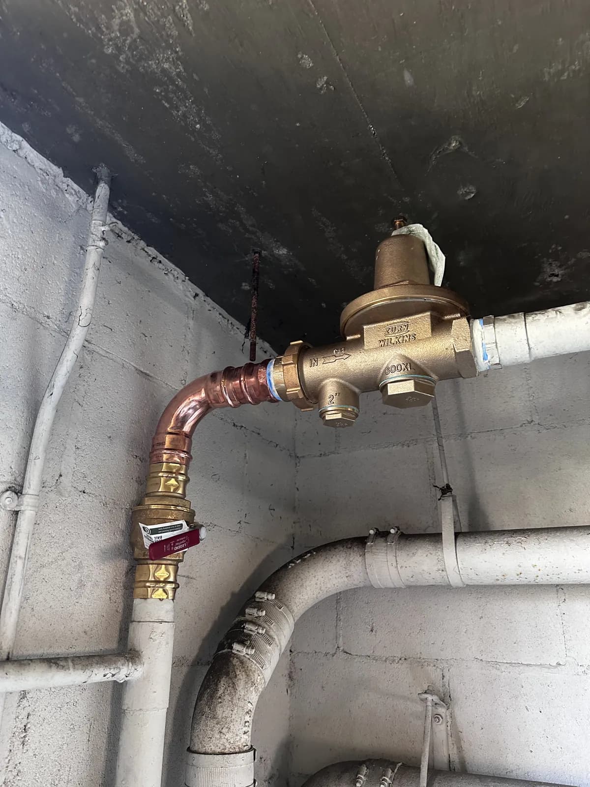 A new brass pressure regulator plumbed on copper with a full-port ball valve below it, against the same block wall