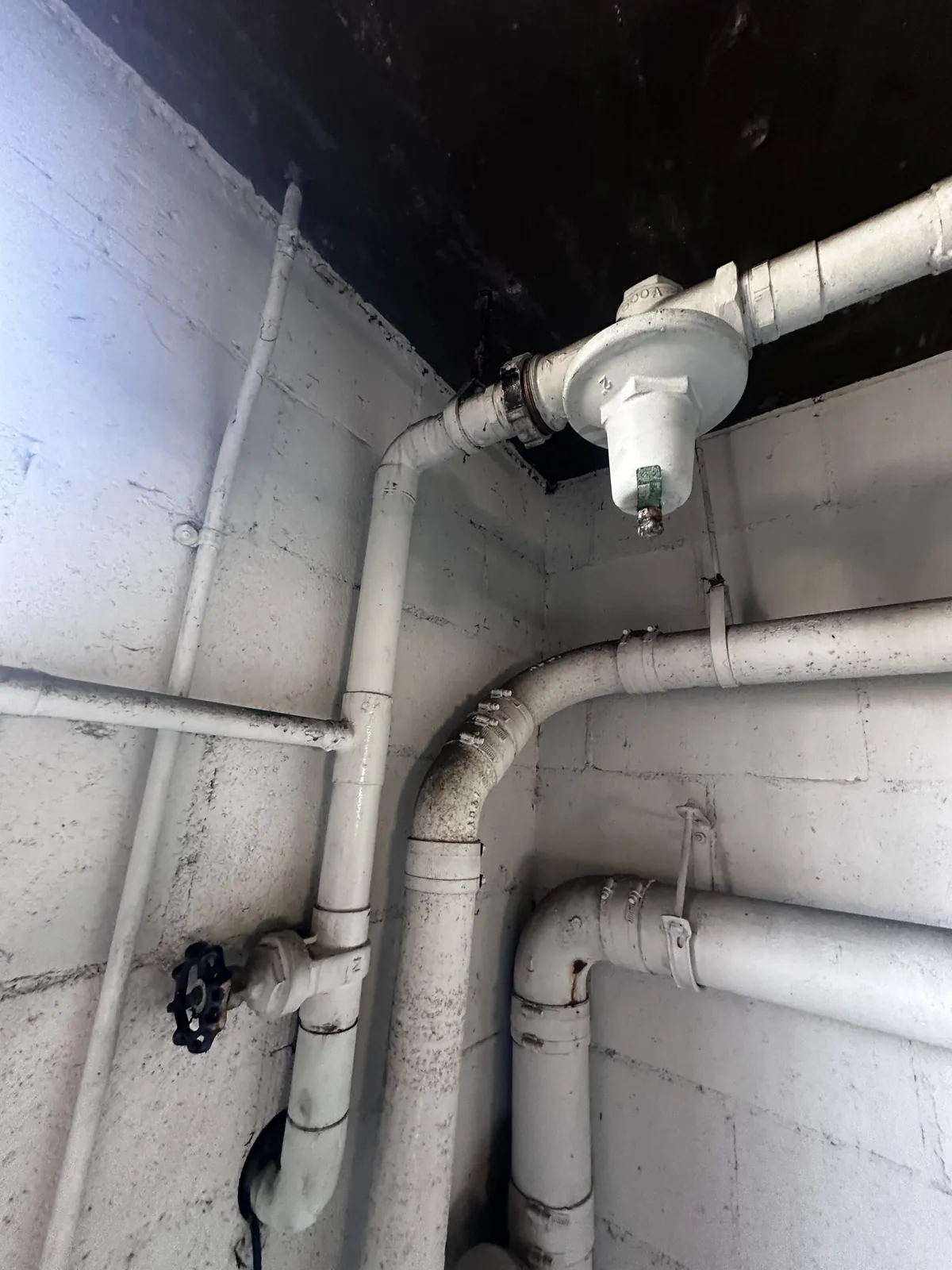 An old water main and pressure regulator on painted galvanized pipe against a block wall, the fittings weathered and a gate valve below