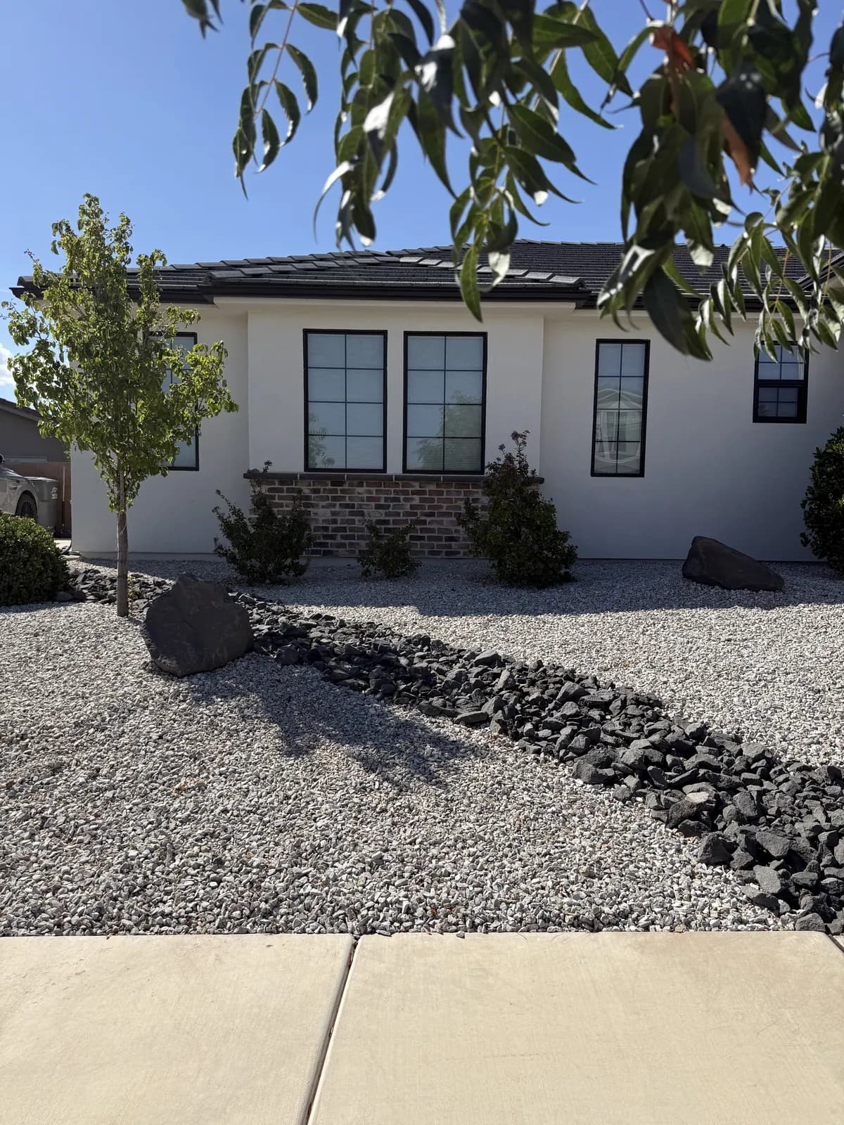 A converted front yard in pale gravel with a dry creek of dark rock running through it, young trees and low desert shrubs along a white stucco house