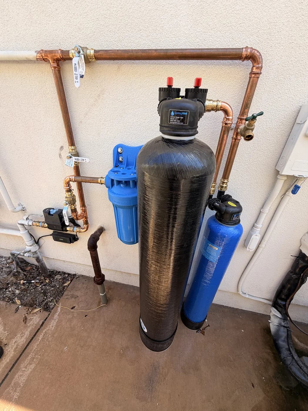 Whole-home water treatment mounted on an exterior stucco wall: a tall carbon tank and a blue brine tank plumbed in new copper, with a blue sediment housing and a bypass loop above